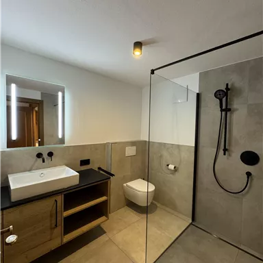 Ein modernes Bad mit Glasdusche und eleganten grauen Fliesen. Der Waschtisch aus Holz ergänzt die schlichte, stilvolle Einrichtung.