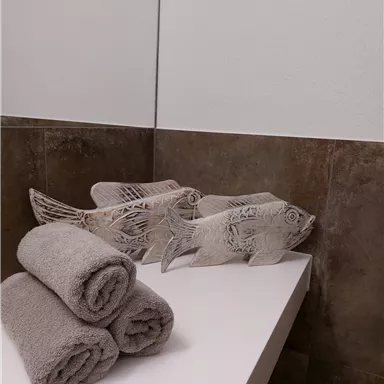 Drei handtuchförmige Fische aus Holz stehen dekorativ auf einem weißen Regal. Darunter liegen mehrere graue Handtücher.