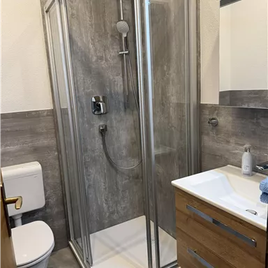 Ein modernes Badezimmer mit einer Glasdusche und Holzwaschtisch. Die Wände sind in neutralen Grautönen gehalten.