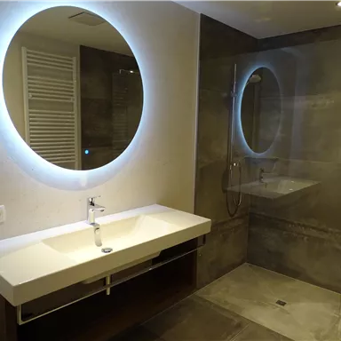 Ein modernes Badezimmer mit zwei runden Spiegeln und einer gläsernen Dusche. Die Wände sind in neutralen Grautönen gehalten und das Waschbecken hat eine schlichte, elegante Form.