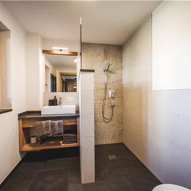 A modern bathroom with an open shower and a wooden shelf. Bright walls and large windows create a friendly atmosphere.