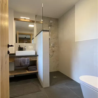 A modern bathroom with gray tiles and an elegant sink. The shower is separated by a glass wall.