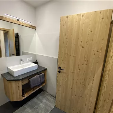 A modern bathroom with wooden paneling and a sink on a gray countertop. In the background is a mirror and an open door leading to another room.