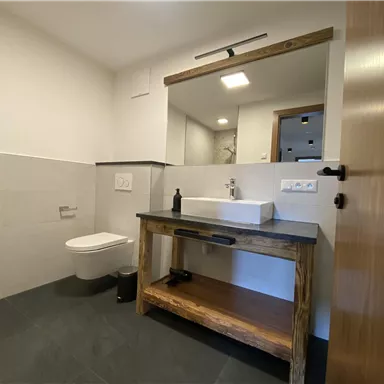 A modern bathroom with a large wooden dresser and a simple sink. The walls are light, and the floor is dark tiled.