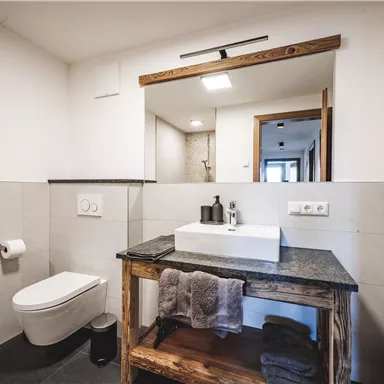 A modern bathroom with a sink on a wooden bench and an elegant toilet area. The walls are designed in light colors and there is plenty of light.