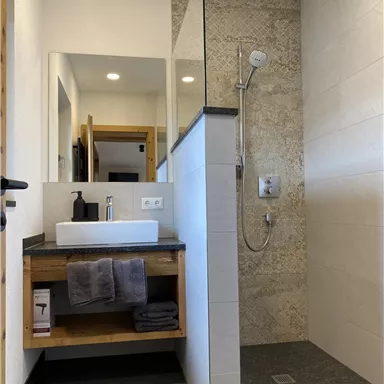 A modern bathroom with a shower and a sink. The walls are tiled in light colors and there are wooden shelves for towels.