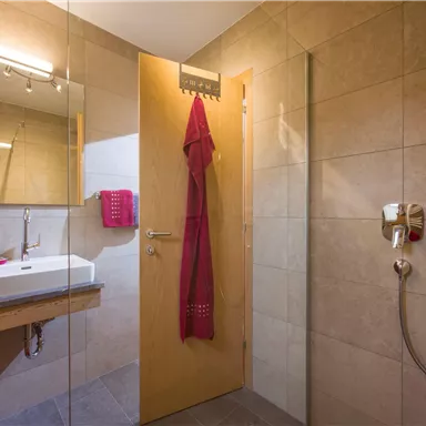 Ein modernes Badezimmer mit einer Duschkabine und einem Waschtisch. An der Wand hängt ein roter Bademantel und es gibt eine große Spiegelfläche.