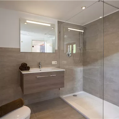 Ein modernes Badezimmer mit einer Glasdusche und hellen Fliesen. Die Ausstattung umfasst ein Waschbecken mit Spiegel und Handtüchern.