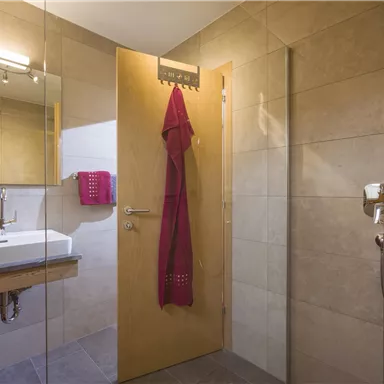 Ein modernes Badezimmer mit Fliesen und einem Holzwaschtisch. An der Wand hängt ein roter Bademantel, und ein großer Spiegel ist sichtbar.