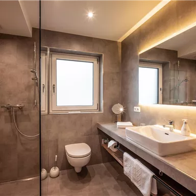 A modern bathroom with a shower and a sink. The walls are painted in neutral gray tones and there is plenty of daylight coming through the window.