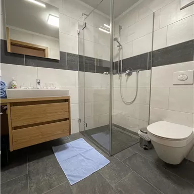 Ein modernes Badezimmer mit einer Glasdusche und einer Holzkommode. Der Boden ist mit dunklen Fliesen ausgelegt und ein blauer Badeteppich liegt auf dem Boden.