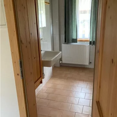 A bright hallway with tiled flooring leads to a bathroom. The bathroom has a window, a radiator, and a sink.