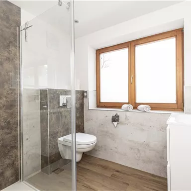Ein modernes Badezimmer mit Dusche, WC und Waschbecken. Große Fenster sorgen für helles Licht und eine angenehme Atmosphäre.