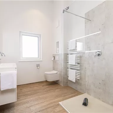 Ein modernes Badezimmer mit einem Waschbecken, einer Dusche und einem Fenster. Der Boden ist aus Holz und es sind frische Handtücher vorhanden.