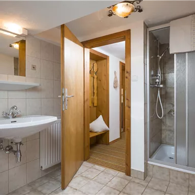 Ein modernes Badezimmer mit Dusche, Waschbecken und Holzdetails. Die Wände sind mit hellen Fliesen gestaltet, und es gibt eine offene Tür zu einem weiteren Raum.