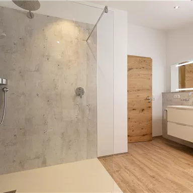 Ein modernes Badezimmer mit einer begehbaren Dusche und einem hellen Holzfußboden. Der Raum ist minimalistisch und gut beleuchtet, mit einem großen Spiegel und eleganten Möbeln.