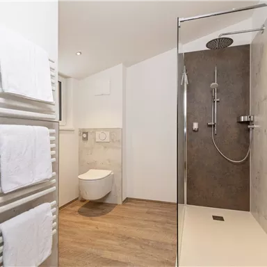 Ein modernes Badezimmer mit einer Dusche und einem WC. Handtücher hängen an einem Heizkörper, und der Boden ist mit Holzoptik gestaltet.