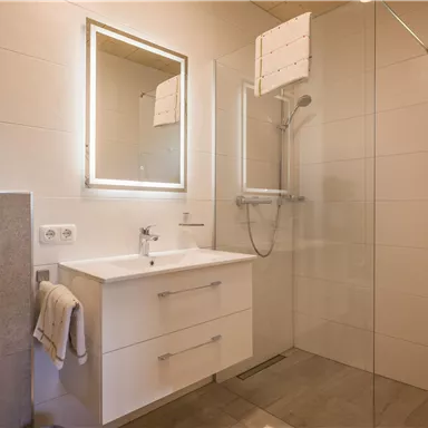 Ein modernes Badezimmer mit einer Dusche und einem großen Spiegel. Die Wände sind hell gefliest und es gibt einen minimalistischen Waschbeckenbereich.