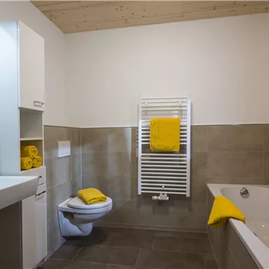 Ein modernes Badezimmer mit einer Badewanne und einem Waschbecken. Gelbe Handtücher und Vorhänge setzen farbige Akzente im Raum.