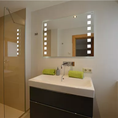 A modern bathroom with a glass shower area and an elegant vanity. The mirror has integrated lights, and green towels add color to the space.