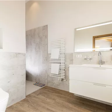 Ein modernes Badezimmer mit hellem, minimalistischen Design. Es gibt eine Dusche, ein Waschbecken und eine angenehme Holzoptik auf dem Boden.