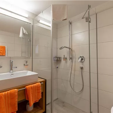 Ein modernes Badezimmer mit einer Dusche und einem Waschbecken. Orangefarbene Handtücher verleihen dem Raum einen farbenfrohen Akzent.