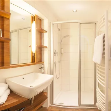 Ein modernes Badezimmer mit einer Dusche, einem Waschbecken und einem Holzregal. Die Wände sind hell gefliest und der Raum wirkt gut beleuchtet.