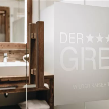 Ein elegantes Badezimmer mit einem modernen Waschbecken und einem vertäfelten Holzdesign. Die gläserne Tür zeigt den Schriftzug „DER GREIL“ in stilvollem Design.