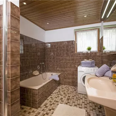 Ein Badezimmer mit braunen Fliesen und Holzdecke. Es enthält eine Badewanne, ein Waschbecken und eine Waschmaschine.