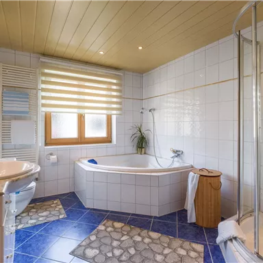 Ein modernes Badezimmer mit einer Badewanne und einer separaten Dusche. Die Wände sind weiß gefliest, und der Boden hat blaue Fliesen mit einem Teppich.