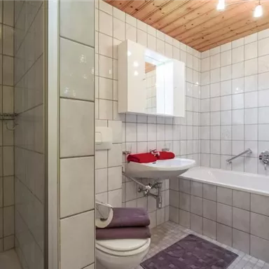 Ein helles Badezimmer mit einer Dusche und einer Badewanne. Die Wände sind gefliest und es gibt Handtücher in Rot und Lila.