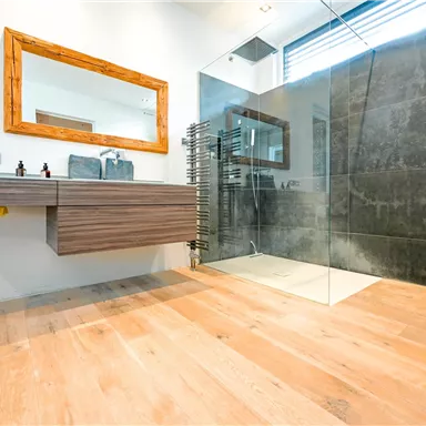 Ein modernes Badezimmer mit einem Holzwaschtisch und einem großen Spiegel. Die Dusche ist durch eine Glaswand abgetrennt, und der Boden ist aus hellen Holzplanken.