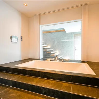 Ein modernes Badezimmer mit einer freistehenden Badewanne auf einer erhöhten Plattform. Große Fenster sorgen für viel natürliches Licht und einen Blick auf eine Treppe.
