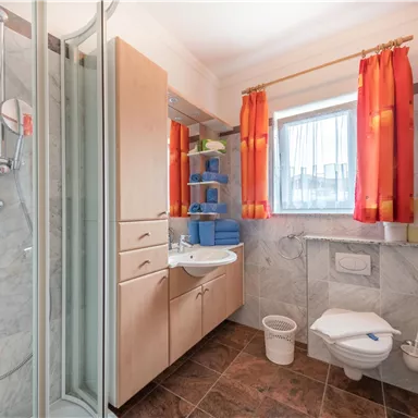 A modern bathroom with a shower, sink, and toilet. The walls are kept in neutral tones, with red curtains and wooden shelves.