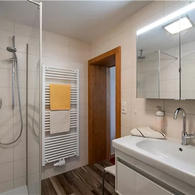 Ein modern gestaltetes Badezimmer mit einer Dusche und einem Waschbecken. Die Wände sind hell gefliest und es gibt Holzdetails im Boden.