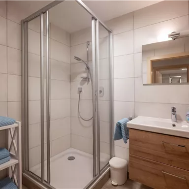 Ein modernes Badezimmer mit einer Dusche und einem Waschbecken. Die Wände sind hell gefliest und es gibt frische Handtücher in blauen Farben.