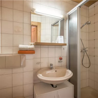 A small bathroom with a shower and a sink. The walls are tiled, and there are some towels on a shelf.