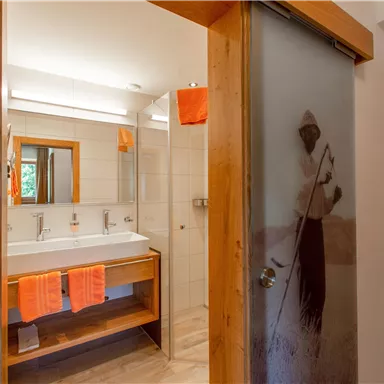 Ein modernes Badezimmer mit Doppelwaschbecken und Holzdetails. Orangefarbene Handtücher hängen stilvoll an der Wand.
