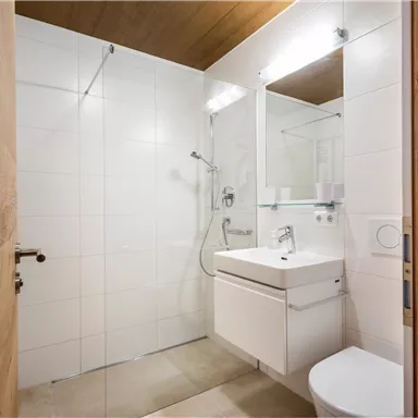 A modern bathroom with a shower, a sink, and a mirror. The walls are light tiled, and there are wooden elements in the room.
