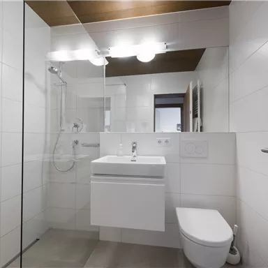 A modern bathroom with a glass shower and a white sink. The walls are light-tiled and there are wood details on the ceiling and door.