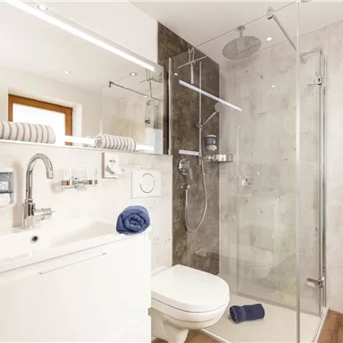 Ein modernes Badezimmer mit einer Dusche und einem Waschbecken. Die Wände sind in neutralen Tönen gehalten und es gibt Handtücher in unterschiedlichen Farben.