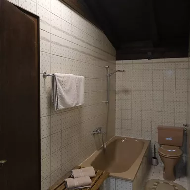 Ein einfaches Badezimmer mit einer Badewanne, einer Toilette und einem Holzregal. Die Wände sind gefliest und die Decke hat Holzdetails.