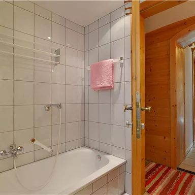 Ein helles Badezimmer mit einer Badewanne und weißen Fliesen. Ein Flur ist im Hintergrund mit Holzdetails zu sehen.