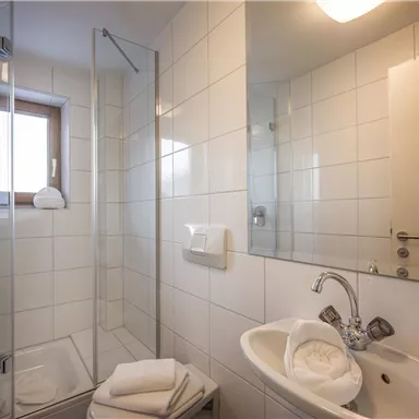 Ein modernes Badezimmer mit einer Glasdusche und einem Waschtisch. Es ist hell und minimalistisch gestaltet, mit weißen Fliesen und einem großen Spiegel.