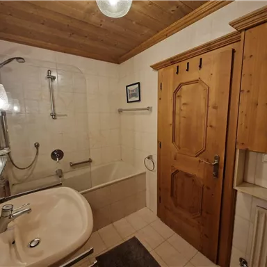 Ein modernes Badezimmer mit einer Badewanne und einer Duschkabine. Die Wände sind hell gefliest und es gibt Holzdetails im Deckenbereich.