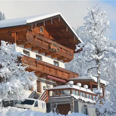 Ein schneebedecktes Gebäude im alpinen Stil umgeben von winterlichen Bäumen. Der Himmel ist klar und die Berge sind im Hintergrund sichtbar.