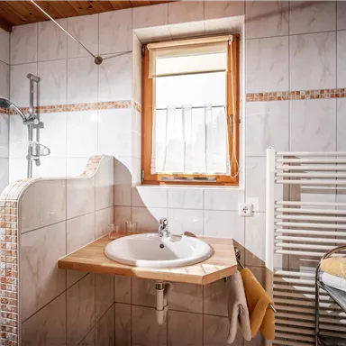 A modern bathroom with white tiles and a sink. There is a shower and a towel radiator, as well as plenty of daylight from the window.