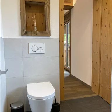 A modern bathroom with a white toilet and a bright wooden door frame. On the wall, there is a wooden shelf with scented sticks.