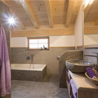 A modern bathroom with a bathtub and a stone sink. The walls are partly clad with an elegant gray material, and there are towels in purple.