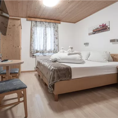 A cozy bedroom with a double bed, a table, and a chair. There are wooden panelings and daylight coming through the window.